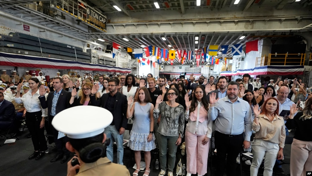 Pessoas levantam a mão direita ao prestarem o Juramento de Fidelidade aos Estados Unidos da América durante uma cerimónia de naturalização a bordo do USS Bataan em 7 de maio de 2024, em Miami.