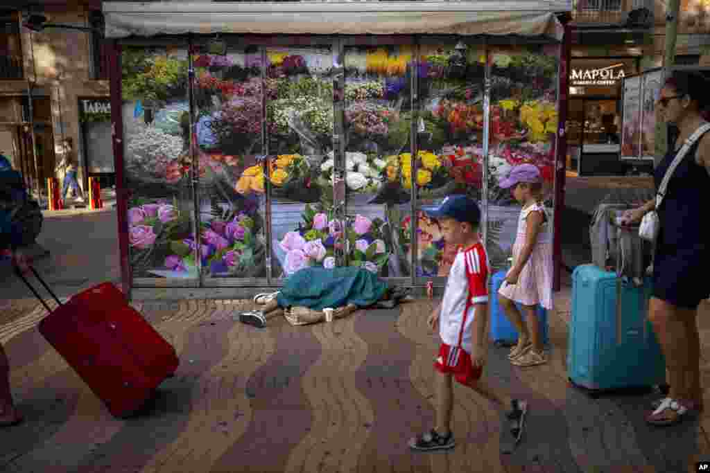 Tourists walk past a homeless man sleeping next to a closed flower shop, along the historic La Rambla promenade of Barcelona, Spain.