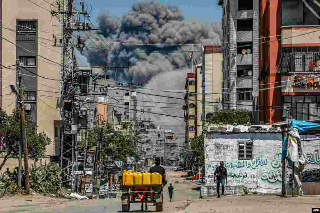 Kepulan asap tebal akibat ledakan tampak di latar belakang saat seorang pria mengendarai gerobak yang ditarik hewan berisi jerigen melintas di kota Nuseirat, Jalur Gaza tengah. (AFP)&nbsp;