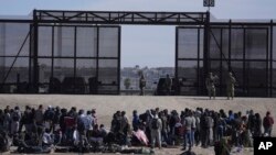 FILE - Migrants who crossed the border from Mexico into the US wait next to the US border wall where US Border Patrol agents stand guard, seen from Ciudad Juarez, Mexico, March 30, 2023. 