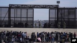 فائل - میکسیکو سے سرحد عبور کر کے امریکہ آنے والے تارکین وطن امریکی سرحدی دیوار کے ساتھ انتظار کر رہے ہیں جہاں امریکی بارڈر پٹرولنگ ایجنٹ محافظ کھڑے ہیں. یہ تصویر 30 مارچ 2023 کو میکسیکو کے سیوڈاڈ واریز سے لی گئی۔فوٹو اے پی