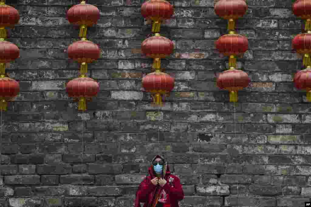 A woman wearing a face mask adjusts her scarf near a wall with lanterns on display in Beijing.