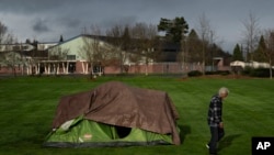 A homeless man walks near a tent with Fruitdale Elementary School in the background at Fruitdale Park in Grants Pass, Oregon, on March 23, 2024. This rural city in southern Oregon may seem like an unlikely poster child for America’s homelessness crisis.