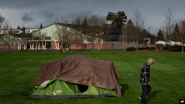 With Fruitdale Elementary School in the background, a homeless person walks near a tent in Fruitdale Park, March 23, 2024, in Grants Pass, Ore. The rural city in southern Oregon has become the unlikely face of the nation's homelessness crisis.