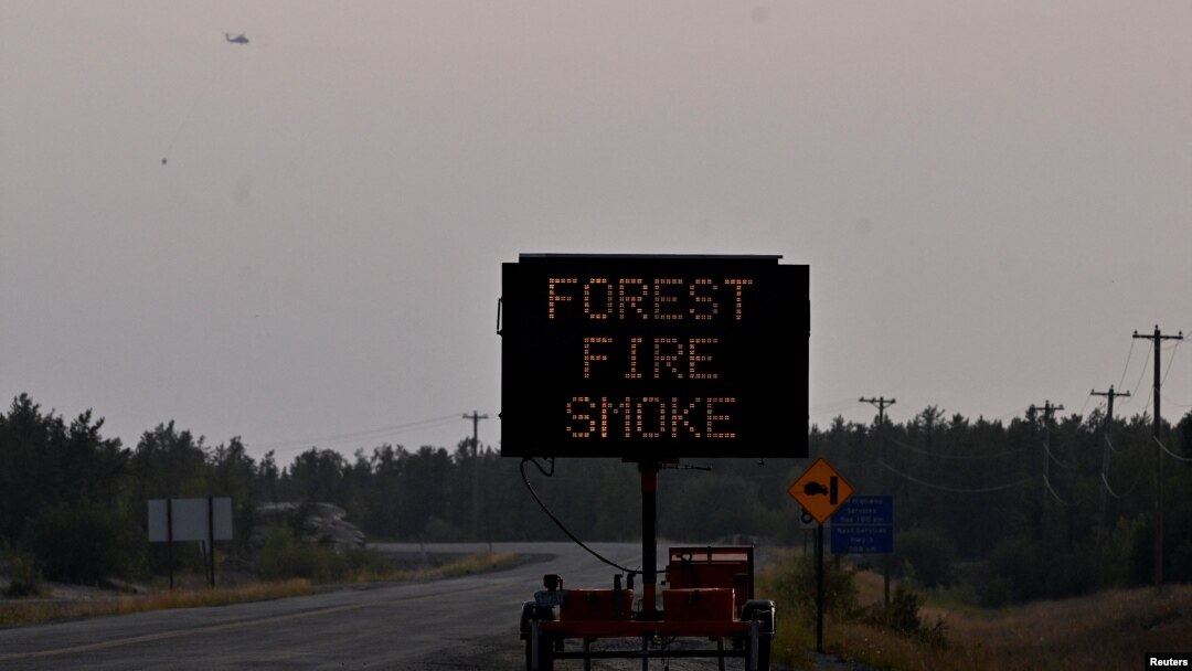 At upper left, a helicopter carrying a water bucket passes a sign warning of smoke as wildfires threatened the Northwest Territories town of Yellowknife, Canada, Aug. 17, 2023.