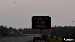At upper left, a helicopter carrying a water bucket passes a sign warning of smoke as wildfires threatened the Northwest Territories town of Yellowknife, Canada, Aug. 17, 2023.