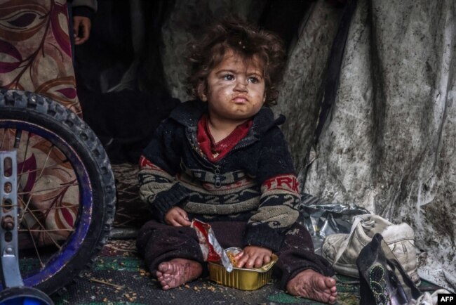 Seorang anak pengungsi Palestina makan makanan dari kotak sambil duduk di tenda darurat di sebuah kamp di samping jalan di Rafah pada 13 Maret 2024, di tengah pertempuran yang sedang berlangsung antara Israel dan Hamas. (Foto: AFP)