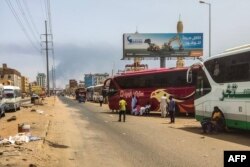 Orang-orang menunggu di samping bus penumpang saat asap mengepul di sebuah daerah di Khartoum tempat pertempuran berlanjut antara tentara Sudan dan pasukan paramiliter, 28 April 2023. (Foto: AFP)