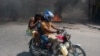 A woman with a child lowers her head as she leaves the Port-au-Prince area on a motorcycle, March 20, 2024. Amid gunfire and a shortage of resources, humanitarians are trying to help as many of the 5.5 million Haitians in need as they can.