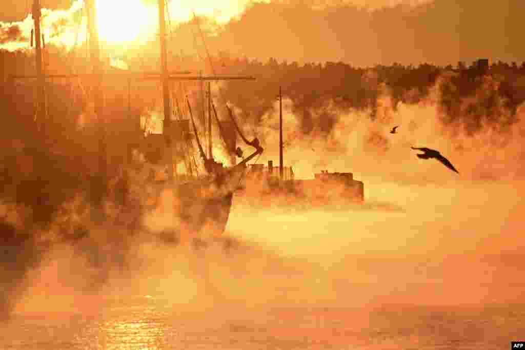 Vapor rises from the water around a historic tri-master charter sailing ship moored in a port of Oslo as morning sun paints the scene in golden light during unusual cold temperatures around -22 degrees Celsius in the Norwegian capital.