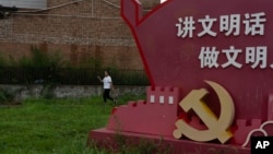 FILE - A woman passes near a government propaganda with the words 'Speak in civilized manners, act in civilized manners' near the emblem for the Communist Party on the outskirts of Beijing, July 25, 2024.