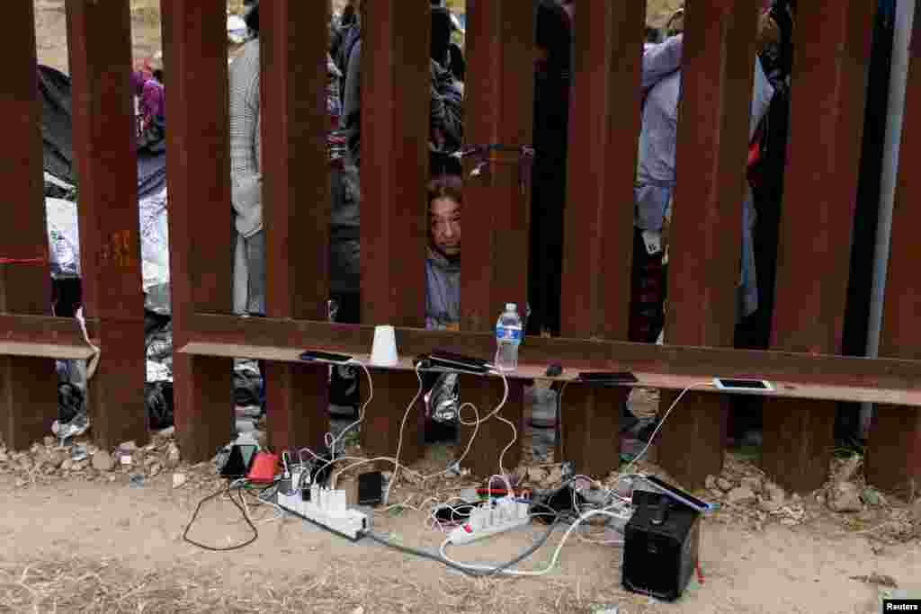 Migrantes cargan sus teléfonos mientras esperan entre las cercas fronterizas.