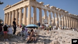 FILE - A tourist drinks water as she and a man sit under an umbrella in front of the Parthenon at the Acropolis during a heat wave in Greece, July 13, 2023.
