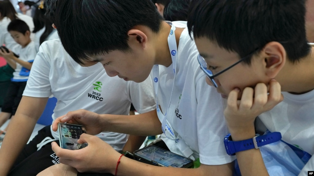 资料照片：2023年8月17日，在北京举行的年度世界机器人大会上，一名学生帮助他的同学通过智能手机玩网络游戏。（美联社照片）(photo:VOA)