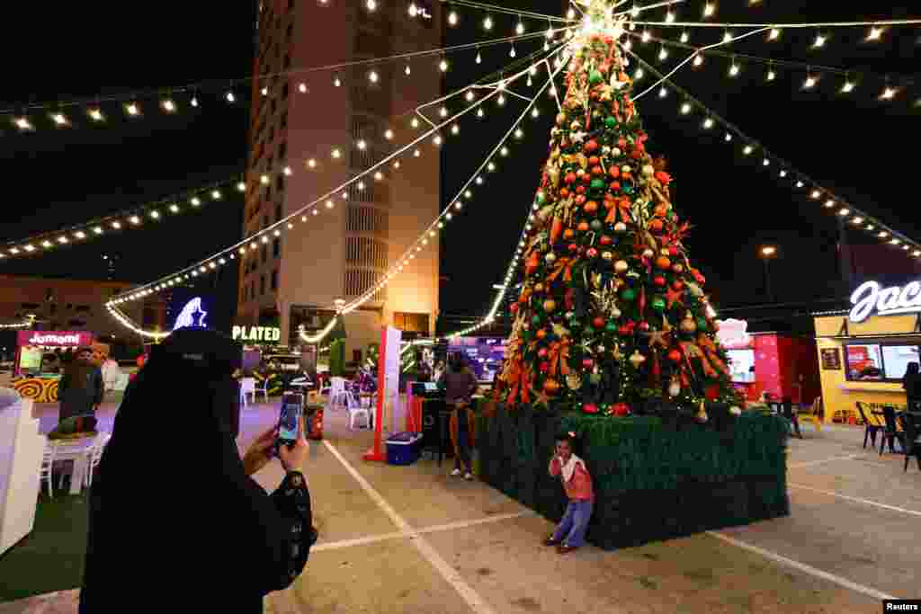 A woman takes a photo of her child next to a Christmas tree at a season festival to celebrate Christmas Eve, in Al A'ali Mall, Manama, Bahrain, Dec. 24, 2023. 