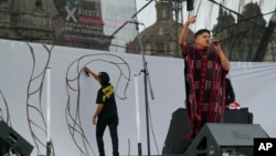 Mexican-Triqui artist Carlos CGH, right, performs during a celebration of Indigenous peoples as artist Alberto Sebastián Bautista Figueroa spray paints a mural on a stage wall at the Zocalo in Mexico City, Aug. 6, 2024.