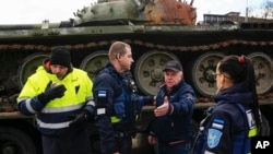 A security guard, left, blows out a candle as police officers talk with a man who placed it next to a destroyed Russian T-72B3 tank installed as a symbol of the Russia-Ukraine war, in Freedom Square in Tallinn, Estonia, March 1, 2023.
