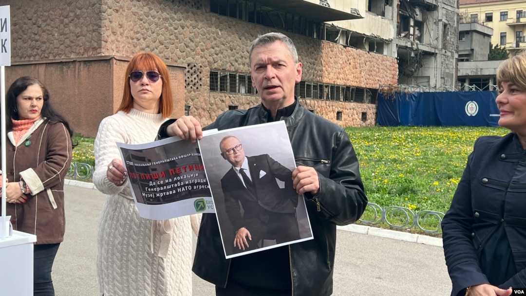 Lider Ekološkog ustanka Aleksandar Jovanović Ćuta pokazuje plakat peticije za podizanje spomen obeležja na mestu uništene zgrade Generalštaba VS i portret funkcionera vladajuće SNS Gorana Vesića, ispred zgrade Generalštaba u Beogradu, 15. marta 2024. (Foto: VoA/Rade Ranković)
