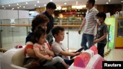 在上海一家商场里一名女子抱着一个小女孩带着一个小男孩驾驶玩具汽车。（2021年6月1日）