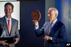 In this image provided by the White House, President Joe Biden holds a wooden presidential seal presented to him by Situation Room director Marc Gustafson, left, in the West Wing of the White House, Sept. 5, 2023.