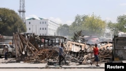 FILE - People walk past remains of vehicles near the presidential palace, after they were set on fire by gangs, as violence spreads and armed gangs expand their control over the capital, in Port-au-Prince, Haiti, March 25, 2024.