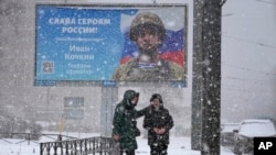 A woman talks to a boy as they walk under a snowfall past a billboard with a portrait of a Russian soldier awarded for action in Ukraine and the words "Glory to the heroes of Russia", in St. Petersburg, Russia, Feb. 10, 2023