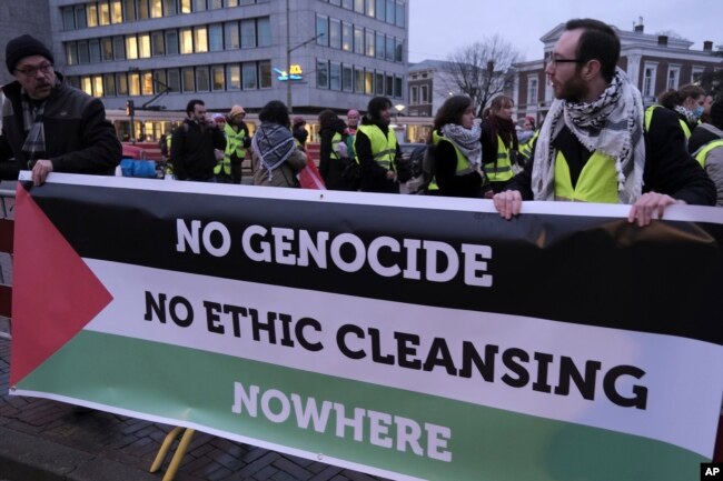 Orang-orang berdiri di depan spanduk di luar Mahkamah Internasional sebelum sidang di Den Haag, Belanda, Jumat, 12 Januari 2024. (AP/Patrick Post)