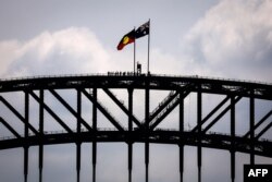 (FILES) Wisatawan berdiri di bawah bendera nasional Aborigin dan Australia yang terletak di atas Jembatan Pelabuhan Sydney pada 27 September 2023, menjelang referendum "Voice". (DAVID GREY / AFP)