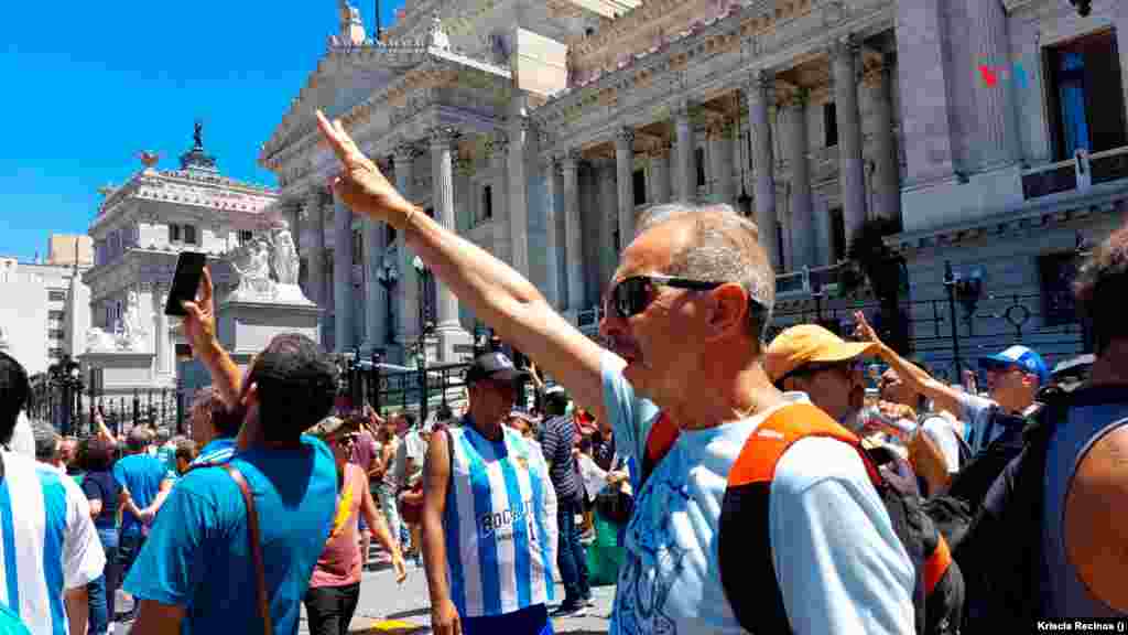 Un hombre levanta su mano y hace la señal peronista, mientras en la plaza se canta el himno nacional de Argentina.