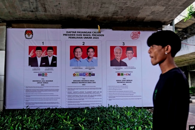 Seorang pria berjalan melewati spanduk KPU yang menampilkan foto calon presiden, Jakarta, Senin, 22 Januari 2024. (AP Photo/Dita Alangkara)