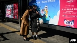 FILE - Two women look at a mobile phone in front of billboards at a bus stop in Beijing, China, Nov. 11, 2023.