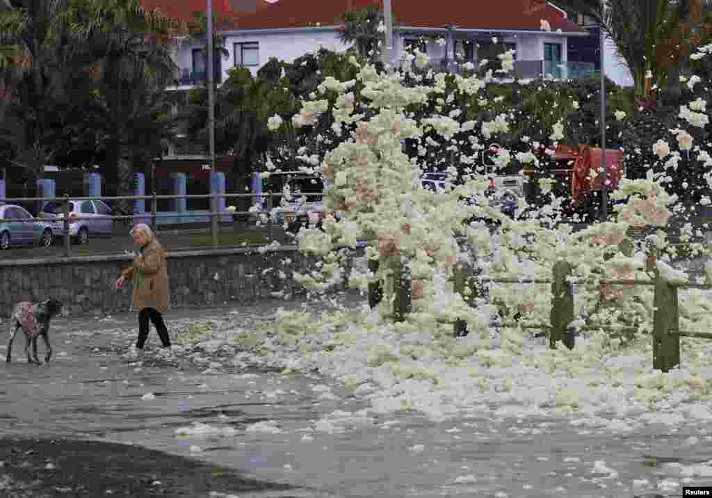 A woman with her dog avoids sea foam at Three Anchor Bay during a cold front in Cape Town, South Africa.