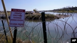 FILE - Migrants cross the Rio Grande into the U.S. from Mexico behind concertina wire and a sign warning that it's dangerous and illegal to cross, Jan. 3, 2024, in Eagle Pass, Texas.