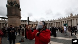 A woman wearing a face mask to curb the spread of COVID-19, uses her phone as Pope Francis recites the Angelus noon prayer from the window of his studio overlooking St.Peter's Square, at the Vatican, March 14, 2021. 