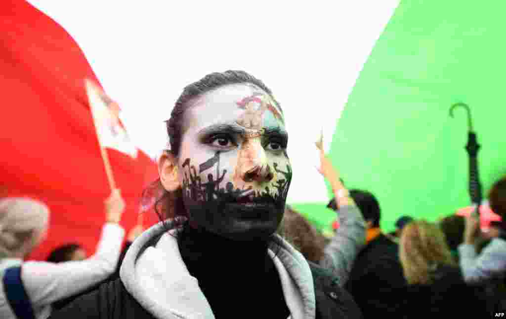 A protester wearing face-paint depicting France&#39;s iconic &quot;Marianne&quot; leading an uprising, stands under a giant Iranian flag as people gather in support of Iranian woman Mahsa Amini during a protest on Place de la Republique in Paris, following her death in custody.