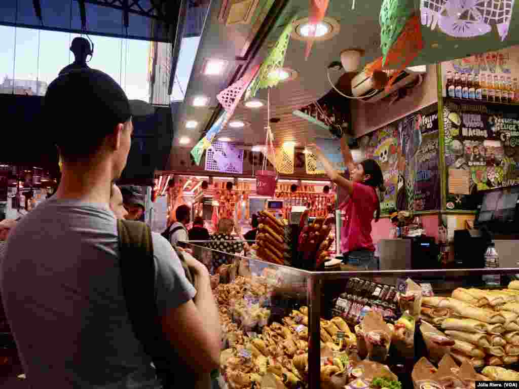 A causa de la vuelta del turismo tras la pandemia y el aumento de migrantes latinoamericanos en Barcelona, cada vez hay más puestos del mercado que ofrecen gastronomía de América Latina. Uno de ellos es Puerto Latino, una tienda que vende productos como arepas, nachos, tequeños y tacos. &nbsp;