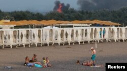FILE - People rest on a beach as smoke and flames rise after explosions at a Russian military airbase, in Novofedorivka, Crimea, Aug. 9, 2022.