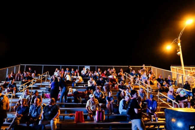 A crowd sits in a bleacher near the Saturn V visitor center several hours before the scheduled NASA moon rocket launch for the Artemis 1 mission to orbit the Moon at the Kennedy Space Center, Monday, Aug. 29, 2022, in Cape Canaveral, Fla. (AP Photo/Brynn