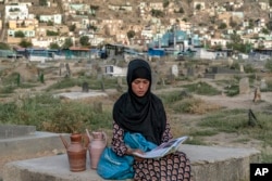 Sara, a 14-year-old, an Afghan girl, sits on a grave and reads a book as she waits for customers to sell water at a cemetery, in Kabul, Afghanistan, Saturday, July 30, 2022. (AP Photo/Ebrahim Noroozi)