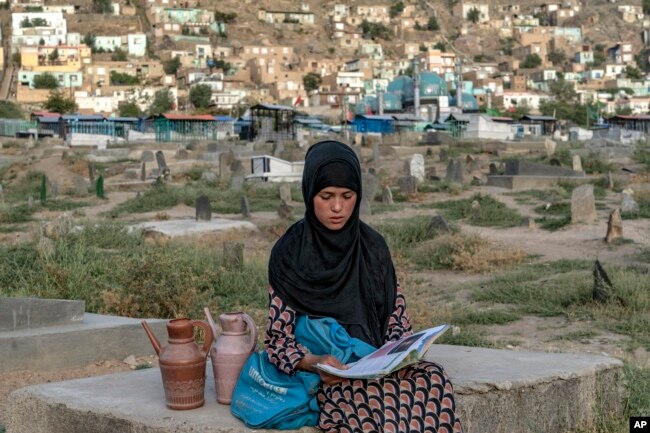 Sara, a 14-year-old, an Afghan girl, sits on a grave and reads a book as she waits for customers to sell water at a cemetery, in Kabul, Afghanistan, Saturday, July 30, 2022. (AP Photo/Ebrahim Noroozi)