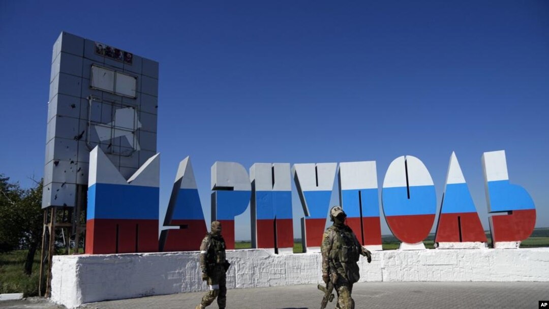 Tentara Rusia berjalan melewati nama kota Mariupol yang dicat ulang dengan warna bendera Rusia di pintu masuk Mariupol, di Donetsk, Ukraina timur, Minggu, 12 Juni 2022. (Foto: AP)