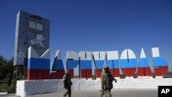 Tentara Rusia berjalan melewati nama kota Mariupol yang dicat ulang dengan warna bendera Rusia di pintu masuk Mariupol, di Donetsk, Ukraina timur, Minggu, 12 Juni 2022. (Foto: AP)