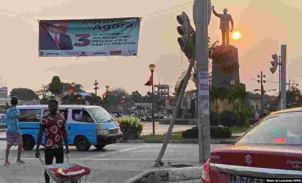 Um jovem atravessa com o carrinho de mão na Praça da Independência. Por trás, um cartaz de campanha da UNITA com a foto do seu líder, Adalberto Costa Júnior. Luanda