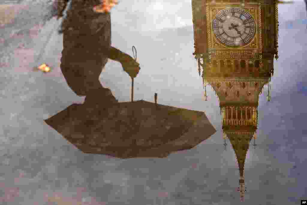 A woman with an umbrella and the Elizabeth Tower, also known as Big Ben, are reflected in a puddle in London.