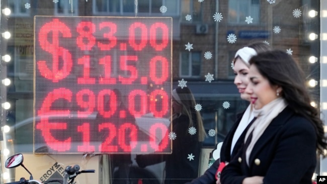 People walk past a currency exchange office screen displaying the exchange rates of U.S. Dollar and Euro to Russian Rubles in Moscow's downtown, Russia, Monday, Feb. 28, 2022. (AP Photo/Pavel Golovkin)