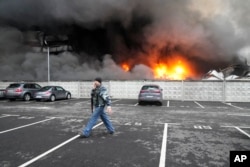 Seorang prajurit Ukraina berjalan melewati saat api dan asap membubung di atas pusat logistik yang rusak setelah penembakan di Kyiv, Ukraina, Kamis, 3 Maret 2022. (Foto: AP)