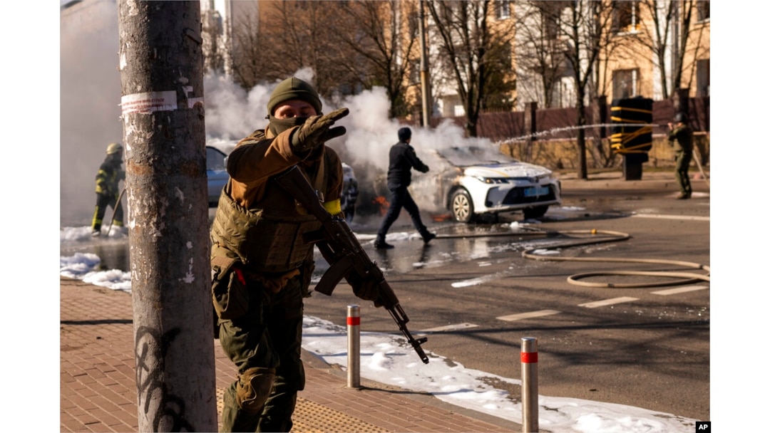 Tentara Ukraina mengambil posisi di luar fasilitas militer saat dua mobil terbakar, di sebuah jalan di Kyiv, Ukraina, Sabtu, 26 Februari 2022. (Foto: AP)