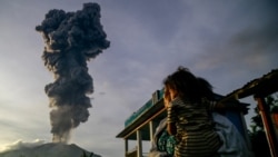 Seorang perempuan dan anaknya menyaksikan kolom abu vulkanis membubung ke udara saat erupsi Gunung Ibu, yang terlihat dari Desa Duono di Kabupaten Halmahera Barat, Provinsi Maluku Utara, Rabu, 15 Januari 2025. (Foto: Azzam/AFP)