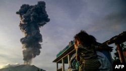 Seorang perempuan dan anaknya menyaksikan kolom abu vulkanis membubung ke udara saat erupsi Gunung Ibu, yang terlihat dari Desa Duono di Kabupaten Halmahera Barat, Provinsi Maluku Utara, Rabu, 15 Januari 2025. (Foto: Azzam/AFP)