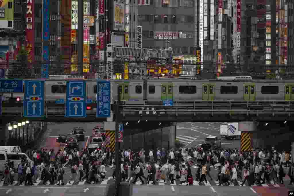 A large crowd passes a pedestrian crossing as a commuter train travels overhead in the Shinjuku district of Tokyo, Japan.